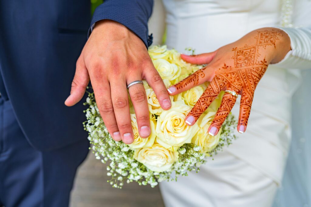Couples' hands with wedding rings on a bouquet of flowers. One hand has Henna designs.