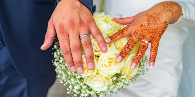 Couples' hands with wedding rings on a bouquet of flowers. One hand has Henna designs.