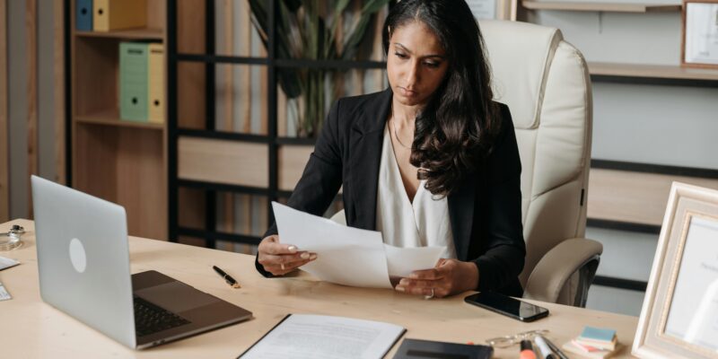 Professional woman reviewing paperwork at an office desk with a laptop, representing business compliance, legal administration and data protection processes.